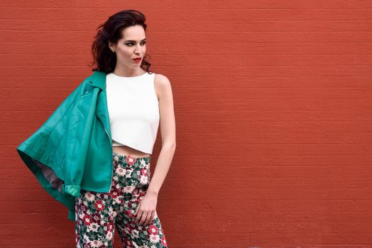 Young brunette woman, model of fashion, wearing green modern jacket and flower pants on red wall. Pretty caucasian girl with long wavy hairstyle. Female with red lips in urban background.