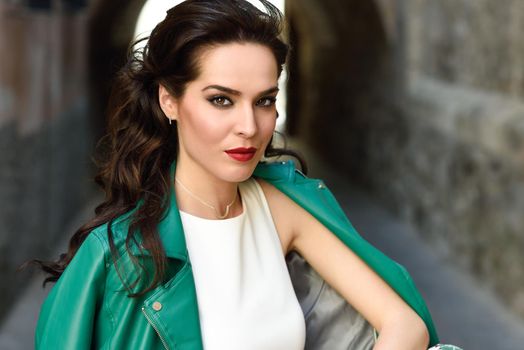 Young brunette woman, model of fashion, wearing green modern jacket. Pretty caucasian girl with long wavy hairstyle. Female with red lips in urban background.