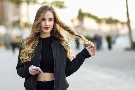 Blonde woman texting with her smart phone in urban background. Beautiful young girl wearing black jacket walking in the street. Pretty russian female with long wavy hair hairstyle.