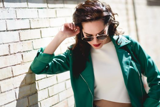 Young brunette woman with aviator sunglasses. Girl, model of fashion, wearing green modern jacket, standing in urban background.