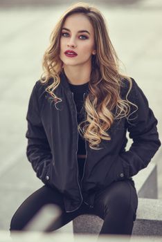 Young blonde girl with beautiful blue eyes wearing black jacket and trousers outdoors. Pretty russian female with long wavy hair hairstyle. Woman in urban background.