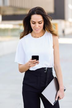 Middle-aged businesswoman working with her smart phone and laptop outdoors. Woman walking near business building with very careful hair.