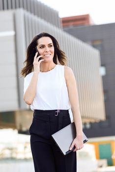 Middle-aged businesswoman working with her smart phone and laptop outdoors. Woman walking near business building with very careful hair.