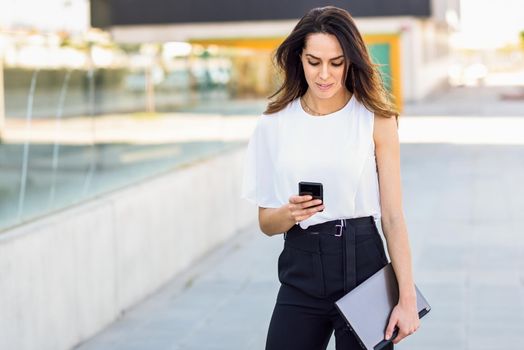 Middle-aged businesswoman working with her smart phone and laptop outdoors. Woman walking near business building with very careful hair.