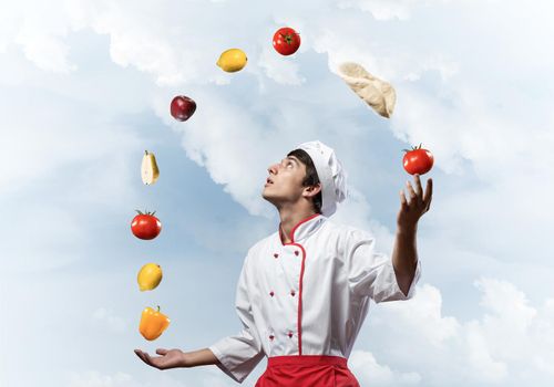 Young male chef juggles with food ingredients. Handsome chef in white hat and red apron on blue sky background. Professional cooking classes advertising. Food retail campaign announcement.
