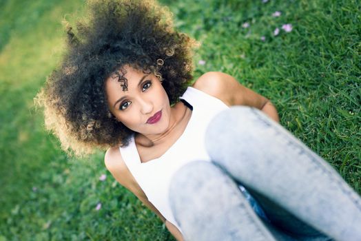 Mixed woman with afro hairstyle laying in urban park. Young black girl wearing casual clothes sitting on the grass looking at camera.