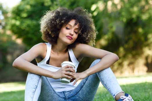 Beautiful young African American woman with afro hairstyle. Girl dreaming with closed eyes with a take away glass in park, sitting on grass wearing casual clothes.