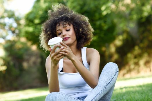 Beautiful young African American woman with afro hairstyle in urban park. Girl reading a take away glass of coffee sitting on grass wearing casual clothes.