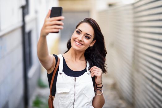 Young woman taking selfie photograph with her smart phone outdoors. Girl wearing denim dress in urban background. Technology concept.