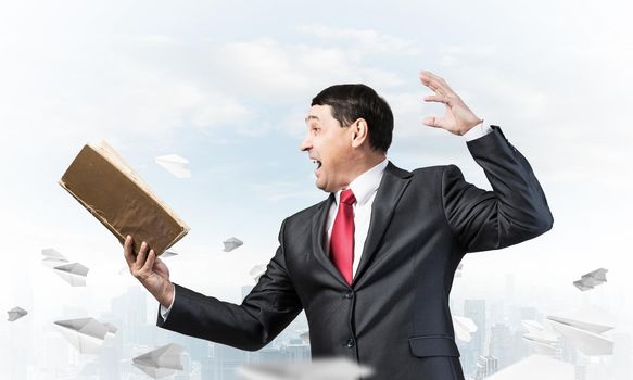 Expressive businessman holding open book. Emotional man in suit and tie standing on downtown background with flying paper planes. Excited manager shouting into book. Professional reference information