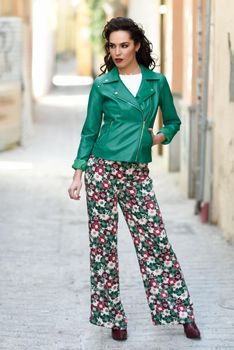 Young brunette woman, model of fashion, wearing green modern jacket and flower pants. Pretty caucasian girl with long wavy hairstyle. Female with red lips in urban background.