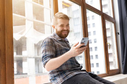 guy hipster with a beard is sitting on the windowsill talking on the video communication using a smartphone and wireless high-speed Internet.