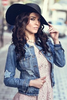 Young brunette woman, model of fashion, wearing denim jacket, hat, and pink dress. Pretty caucasian girl with long wavy hairstyle in urban background.