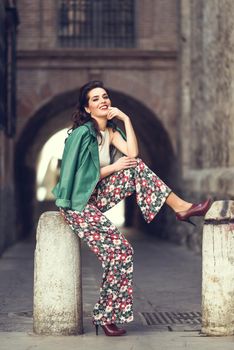 Young brunette woman, model of fashion, wearing green modern jacket and flower pants. Pretty caucasian girl with long wavy hairstyle smiling. Female with red lips in urban background.