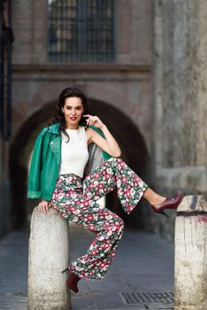 Young brunette woman, model of fashion, wearing green modern jacket and flower pants. Pretty caucasian girl with long wavy hairstyle smiling. Female with red lips in urban background.