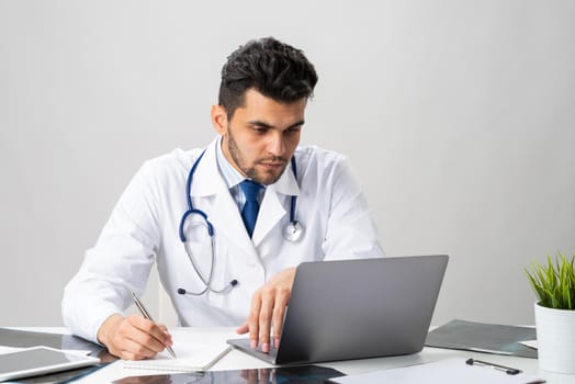 Handsome doctor sitting in office. Young practitioner in white coat with stethoscope at desk with laptop. Professional medical consultation and treatment in modern clinic. Medical healthcare concept
