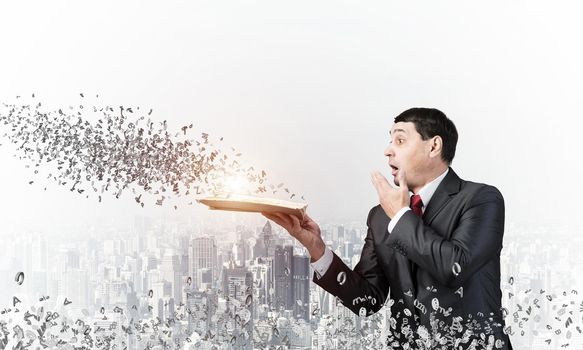 Embarrassed businessman covering mouth with hand and holding open book. Startled man in business suit and tie standing on city background with flying various letters. Business education and knowledges