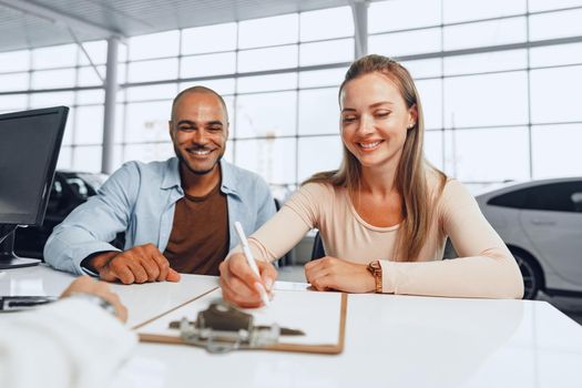 Beautiful young couple signs documents at car dealership showroom sitting at the table