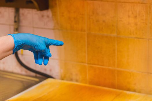 Woman's housewife hand in a household glove points to a dirty sticky coating on the gas stove cover on the kitchen background.