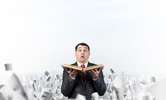 Surprised businessman holding book with open mouth. Joyful adult man in business suit and tie standing on cloudy cityscape background with falling paper documents. Business education and knowledges.