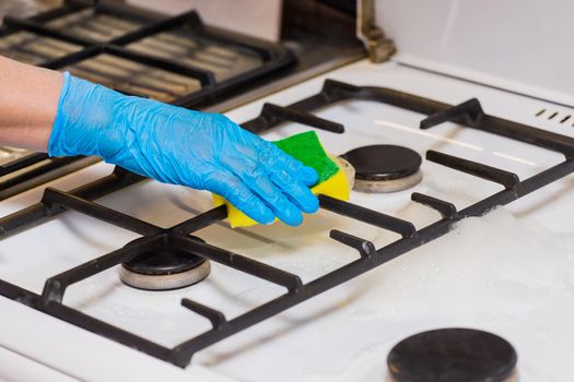 Hand of a woman in a household rubber glove sponges the grate of a gas stove and burners on kitchen, close up.