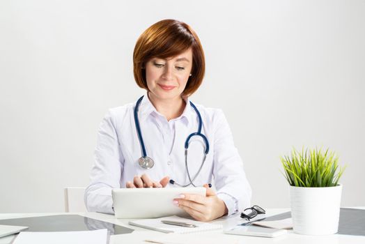Beautiful doctor using tablet computer in office. Family physician in white coat with stethoscope sitting at desk. Professional medical diagnosis and treatment in clinic. Healthcare and medicine.
