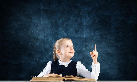 Smiling little girl sitting at desk with open book. Research and education in school. Happy schoolgirl in school wear finger pointing up. Clever girl reading book on deep blue plexus background