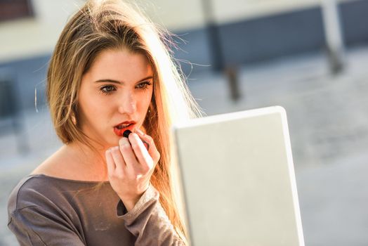 Young woman applying lipstick looking at tablet in urban background