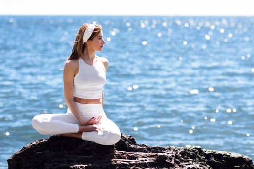 Young woman doing yoga in the beach. Female wearing white sport clothes in lotus figure with defocused sea background.