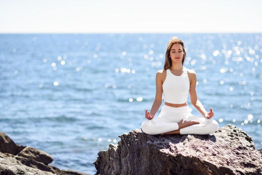 Young woman doing yoga in the beach. Female wearing sport white clothes in lotus figure with defocused background.