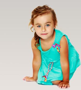 Delicate little blonde girl, dressed in a blue dress standing on her knees. She looks directly into the camera.On a gray background.