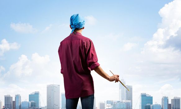 Young male painter artist holding paintbrush. Back view of painter in shirt and bandana on background cloudy sky and cityscape. Creative hobby and artistic occupation. Art school courses concept.