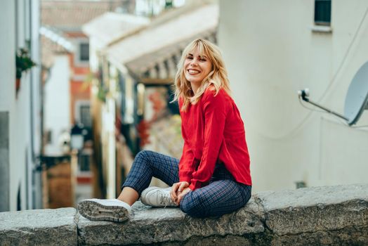 Happy young blond woman sitting on urban background. Smiling blonde girl with red shirt enjoying life outdoors.
