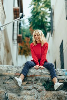 Happy young blond woman sitting on urban background. Smiling blonde girl with red shirt enjoying life outdoors.