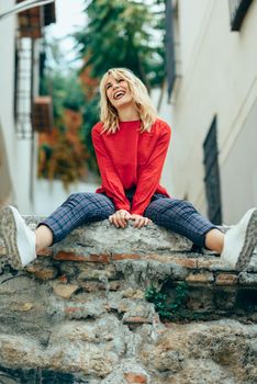 Happy young blond woman sitting on urban background. Smiling blonde girl with red shirt enjoying life outdoors.