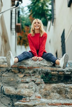 Happy young blond woman sitting on urban background. Smiling blonde girl with red shirt enjoying life outdoors.