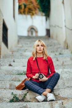 Attractive young blond woman taking photographs with an old slr camera in a beautiful city. Blonde happy woman sitting on urban steps.