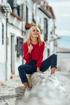Happy young blond woman sitting on urban background. Laughing blonde girl with red shirt enjoying life outdoors.