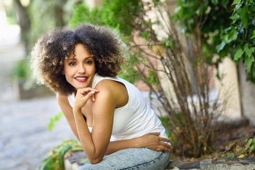 Mixed girl with afro hairstyle sitting in urban park. Young black woman wearing casual clothes.