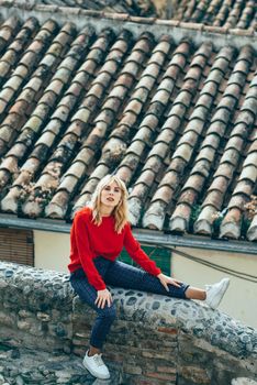 Young blond woman sitting near beautiful roofs of charming old houses.. Smiling blonde girl with red shirt enjoying life outdoors.