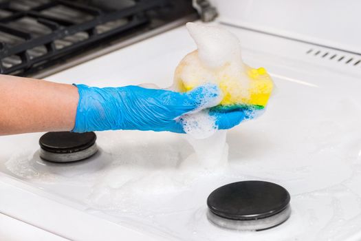 Woman's hand in a household glove washing sponge with foam a dirty gas stove background, close up.