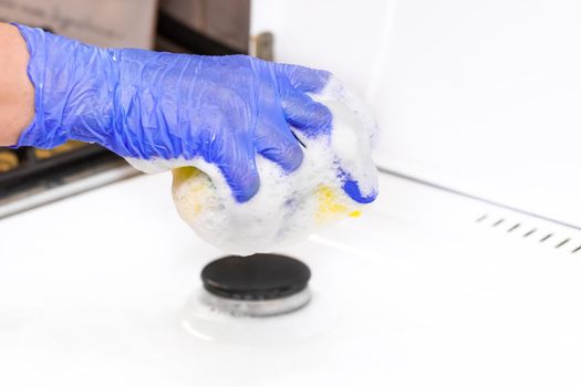 The hand of a housewife in a rubber household glove holds a sponge with foam over the gas stove and is engaged in home cleaning kitchen, close-up.