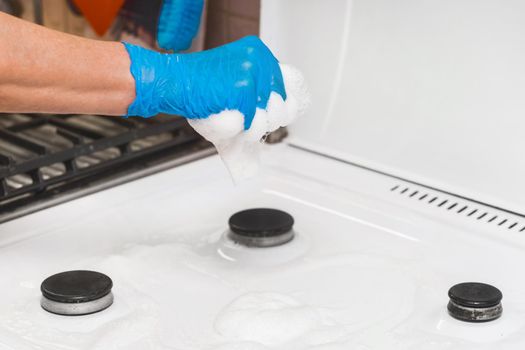 The hand of a housewife girl in a latex household glove squeezes a sponge with foam over a gas stove and burners in the kitchen, close-up.