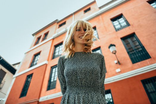 Happy young woman with moving hair in urban background. Blond girl, wavy hairstyle, wearing casual clothes in the street.