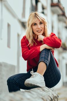 Happy young blond woman sitting on urban background. Smiling blonde girl with red shirt enjoying life outdoors.