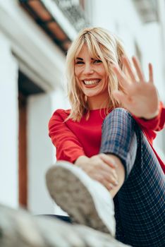 Happy young blond woman sitting on urban background. putting her hand near the camera.