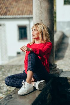 Happy young blond woman sitting on urban background sunbathing in autumn. Smiling blonde girl with red shirt enjoying life outdoors.