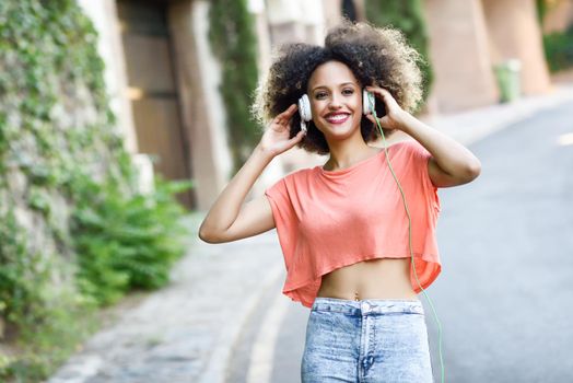 Portrait of young attractive black girl in urban background listening to the music with headphones. Woman wearing orange t-shirt and blue jeans with afro hairstyle