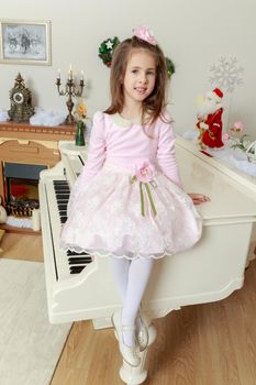 Cute little girl in beautiful pink dress and a rose in her hair.She sat down on the white piano.