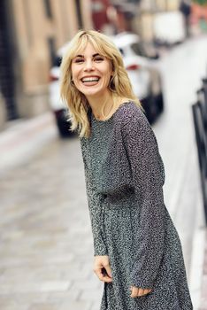 Funny young blonde woman standing on urban background. Smiling blonde girl wearing dress dancing outdoors.